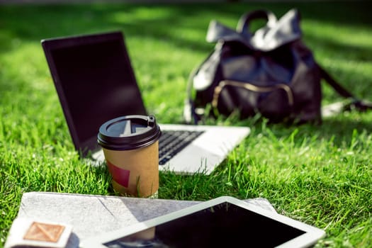Laptop computer on green grass with coffee cup, bag and tablet in outdoor park. Copy space. Still life
