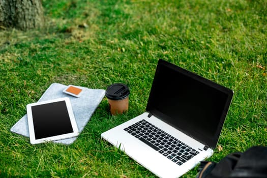 Laptop computer on green grass with coffee cup, bag and tablet in outdoor park. Copy space. Still life