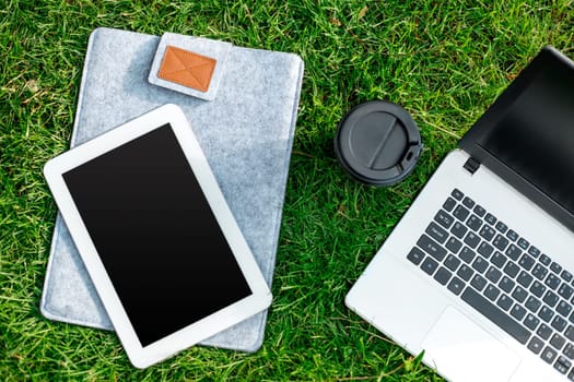 Laptop computer on green grass with coffee cup and tablet in outdoor park. Copy space. Still life