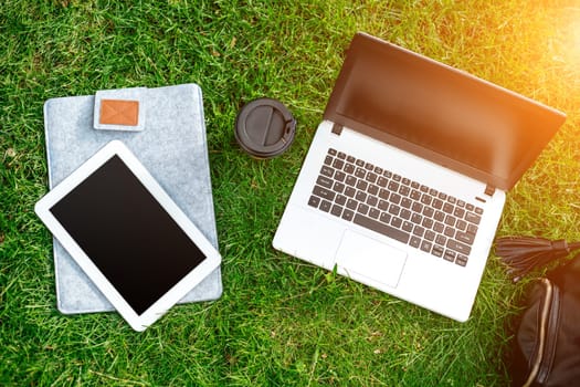 Laptop computer on green grass with coffee cup, bag and tablet in outdoor park. Copy space. Still life. Sun flare