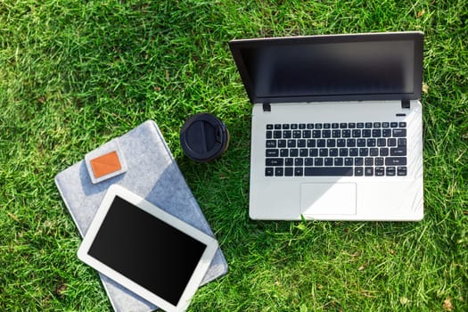 Laptop computer on green grass with coffee cup and tablet in outdoor park. Copy space. Still life