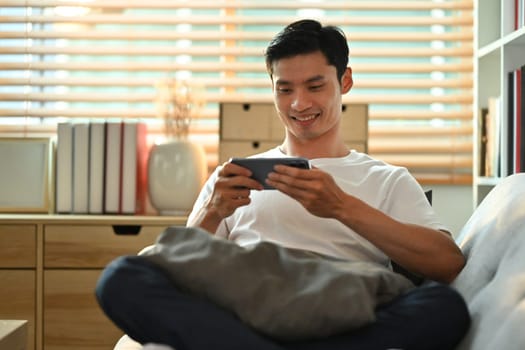 Relaxed asian man in casual wears watching video or playing game on smart phone, sitting on couch at home.
