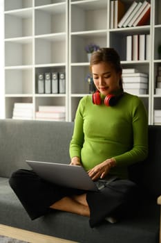 Full length of pretty young woman using laptop computer on couch at home. Freelance, distance, education, e-learning concept.