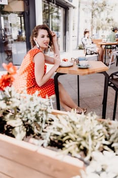 Charming woman in a restaurant, cafe on the street. She sits at the table and eats a cake with a fork. Dressed in a red sundress with white polka dots