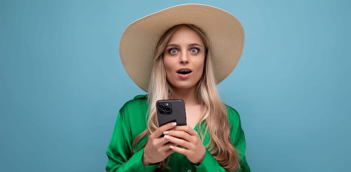 surprised charming young adult woman in a summer hat with a phone in her hands on a blue background with empty space.