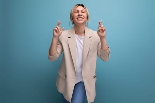 hopeful blonde young adult in beige jacket with crossed fingers on studio background with copy space.