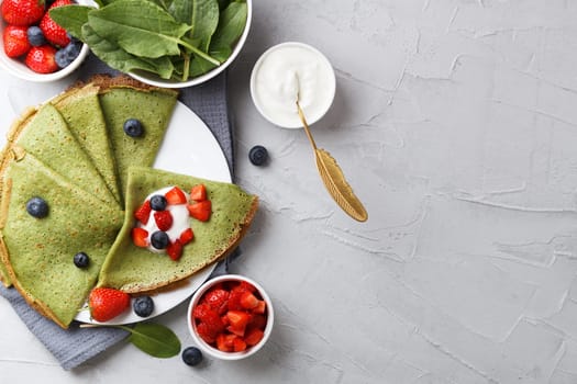 Green pancakes with spinach on a plate with fresh berries and sour cream, on a gray background with textiles. copy space