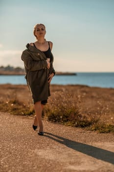 Portrait blonde sea cape. A calm young blonde in an unbuttoned khaki raincoat walks along the seashore, under a raincoat a black skirt and top.