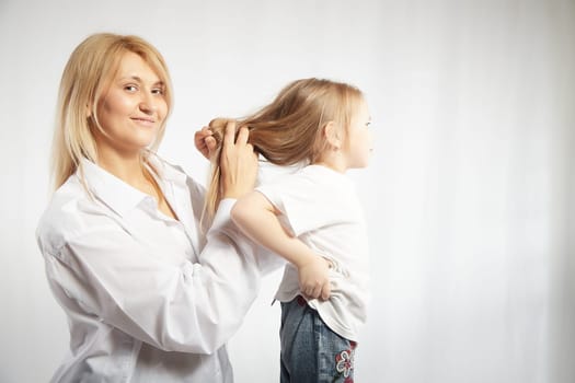 Close portrait of blonde mother and daughter where mom braids hair and makes ponytail and hairstyle for her girl on white background in studio. The concept of love, friendship, caring in family