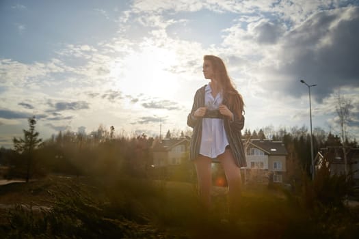 Beautiful girl with long hair in short skirt, white shirt and jacket in village or small town. Tall young slender woman and houses and sky with clouds on background on autumn, spring or summer day