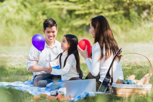 Happy Asian young family father, mother and child little girl having fun and enjoying outdoor sitting on picnic blanket playing balloons at summer garden spring park, Family relaxation concept