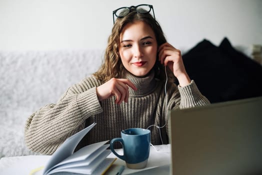 Stylish woman enjoying online education with a cup of coffee at the kitchen table, wearing headphones and writing notes while watching a webinar on her laptop.