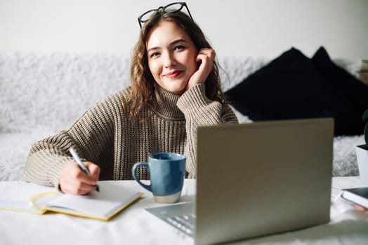 Happy student studying at home, doing homework and preparing for exams in front of laptop, sitting at kitchen table with pen and notebook, smiling at camera.