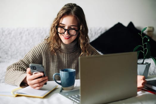SMM specialist working from home at kitchen table, using smartphone to select blogger for product promotion in social media, in front of laptop. Influencer marketing concept.
