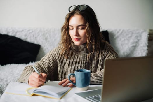 Portrait of a female freelance translator working from home, writing translation in notebook at kitchen table, focused on laptop screen, translating article for website, remote work lifestyle.