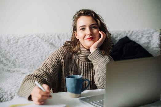 A female PR manager creates an online store content plan from home, working at the kitchen table with a laptop, notebook, and coffee, jotting down notes, while engaging with the camera.