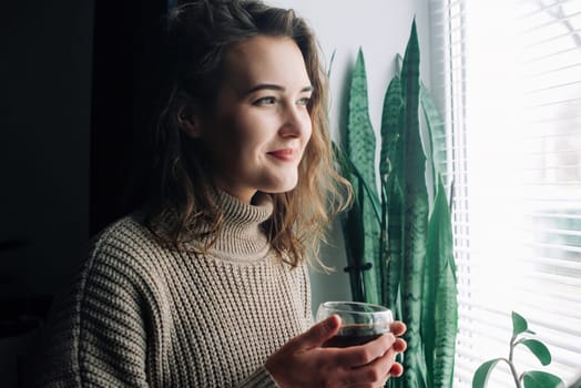 A serene young woman savors a hot cup of tea while admiring the stunning cityscape from a window, taking a peaceful moment to herself to unwind and appreciate the beauty around her