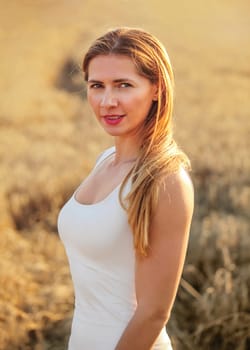 Young brunette woman in white dress, standing in wheat field lit by afternoon sun.