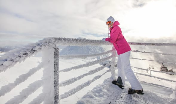 Young woman in pink ski jacket, boots gloves hat and goggles leaning on crystal snow covered fence rail, looking back. Sun shining on top of the mountain piste behind.