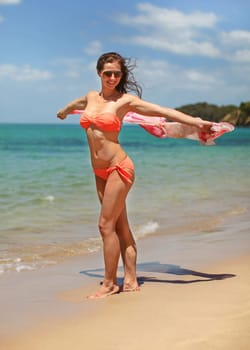 Young sporty woman in orange bikini swimwear and sunglasses, walking on beach, holding scarf behind her that is waving in wind.