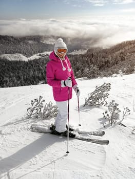 Young woman in pink jacket, skis, ski poles, goggles and hat, posing on the snow covered piste, sun shining to forest behind her.