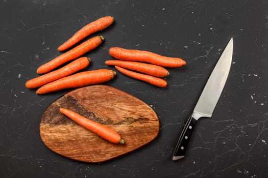 Top down view, orange carrot, chopping board and chef knife next to it, on black marble board.