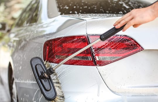 Rear light and back of silver car washed in self serve manual carwash. Brush cleaning shampoo covered surface.