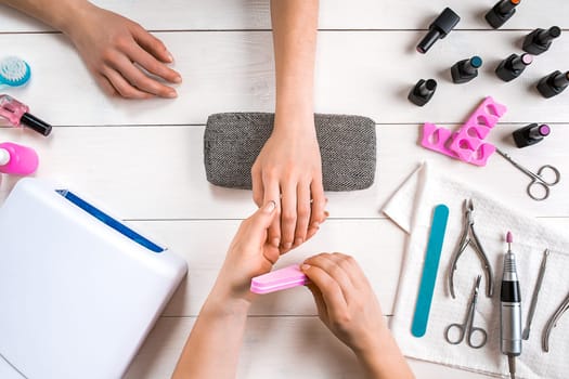 Nail care. Closeup of female hands filing nails with professional nail file in beauty nail salon. Close-up of beautician hands doing perfect manicure on woman's hands. Nail hygiene. Top view