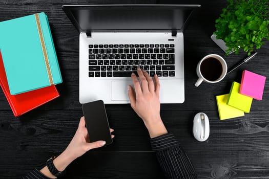 Female hands holding a smart and typing on the keyboard of a laptop on black wooden table. Mock-up with laptop, cup and notebook