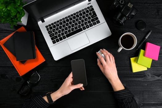 Female hands holding a smart and typing on the keyboard of a laptop on black wooden table. Mock-up with laptop, cup and notebook
