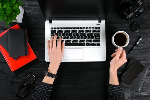 Mix of office supplies and gadgets on a wooden desk background. View from above. Female hands on the laptop keyboard