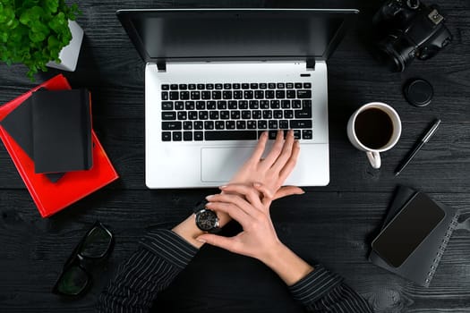 Mix of office supplies and gadgets on a wooden desk background. View from above. Female hands on the laptop keyboard