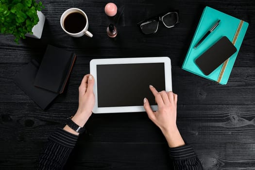 Hand use white tablet on desk table. Top view of human hands, tablet, a cup of coffee, smartphone, notebook and a flower on a wooden table background.