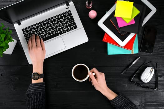 Female hands holding a cup of coffee and typing on the keyboard of a laptop on black wooden table. Mock-up with laptop, cup and notebook