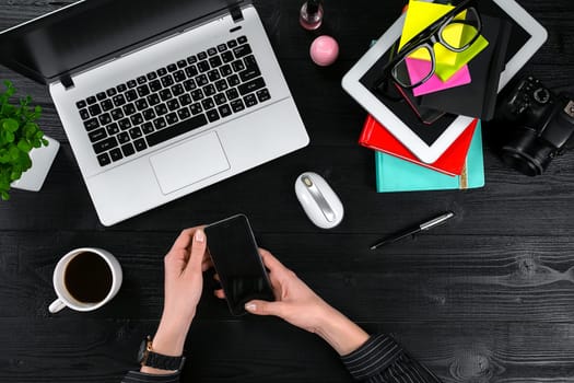 Female hands holding a smart and typing on the keyboard of a laptop on black wooden table. Mock-up with laptop, cup and notebook