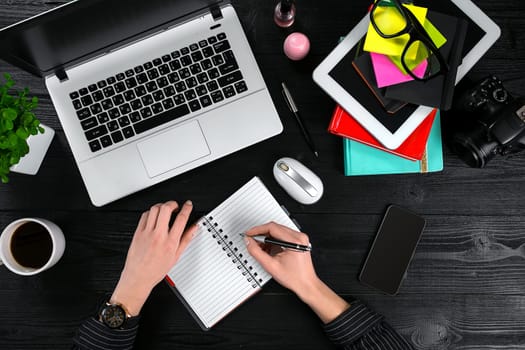 Mix of office supplies and gadgets on a wooden desk background. View from above. Female hands on the laptop keyboard