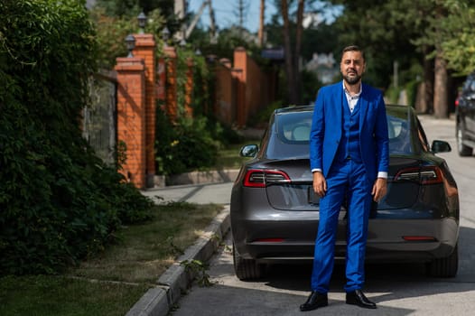 Caucasian bearded man in a blue suit stands near a black car in the countryside in summer