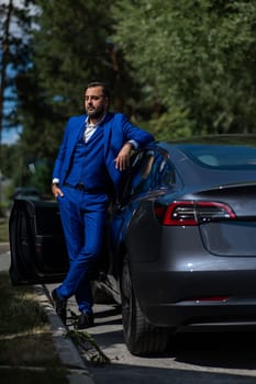 Caucasian bearded man in a blue suit gets out of a black electro car in the countryside in summer