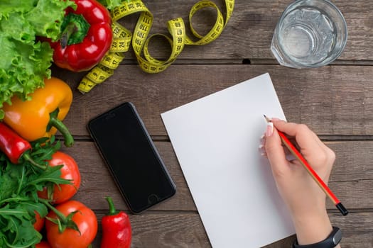 Concept diet, slimming plan with vegetables top view mock up. A female hand in a sports bracelet writes an article about proper nutrition
