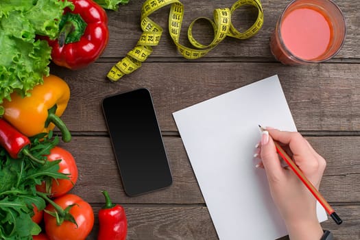 Concept diet, slimming plan with vegetables top view mock up. A female hand in a sports bracelet writes an article about proper nutrition