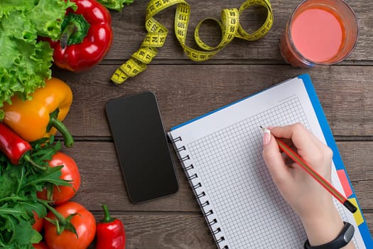 oncept diet, slimming plan with vegetables top view mock up. A female hand in a sports bracelet writes an article about proper nutrition