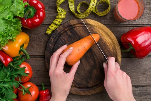 Female hands cutting carrot at table, top view. On the table leaves of lettuce, pepper, a glass of tomato juice, a wooden board and a knife