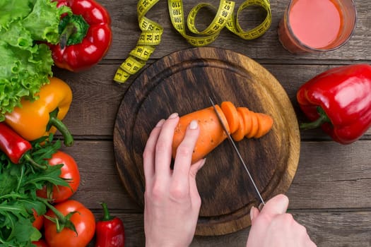 Female hands cutting carrot at table, top view. On the table leaves of lettuce, pepper, a glass of tomato juice, a wooden board and a knife