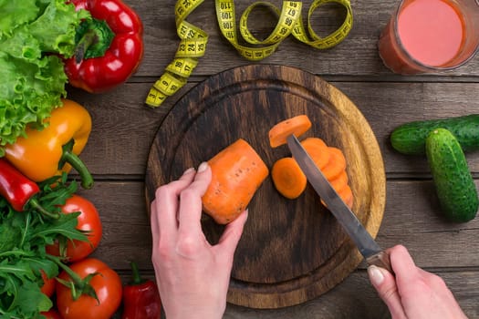 Female hands cutting carrot at table, top view. On the table leaves of lettuce, pepper, a glass of tomato juice, a wooden board and a knife