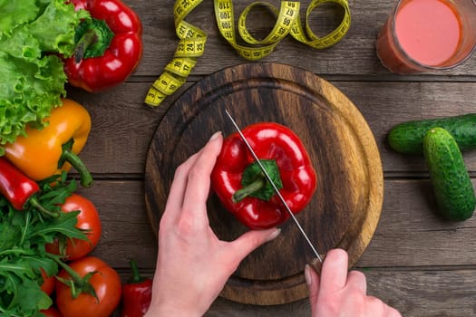 Female hands cutting pepper at table, top view. On the table leaves of lettuce, pepper, a glass of tomato juice, a wooden board and a knife