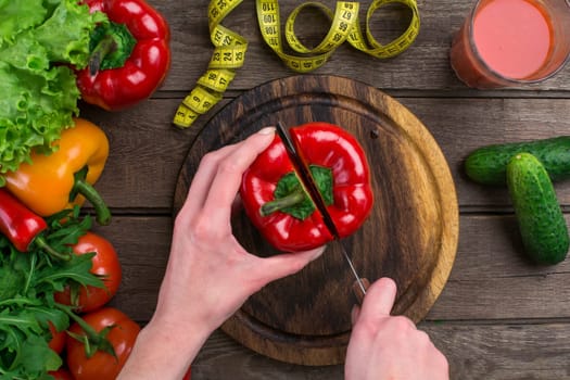 Female hands cutting pepper at table, top view. On the table leaves of lettuce, pepper, a glass of tomato juice, a wooden board and a knife