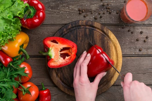 Female hands cutting pepper at table, top view. On the table leaves of lettuce, pepper, a glass of tomato juice, a wooden board and a knife