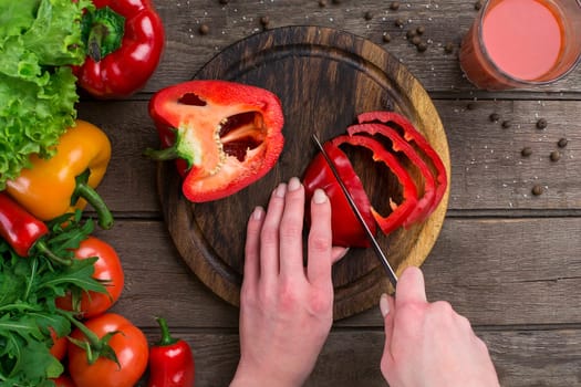 Female hands cutting pepper at table, top view. On the table leaves of lettuce, pepper, a glass of tomato juice, a wooden board and a knife