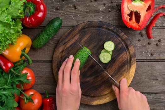 Female hands cutting cucumber at table, top view. On the table vegetables and a wooden board