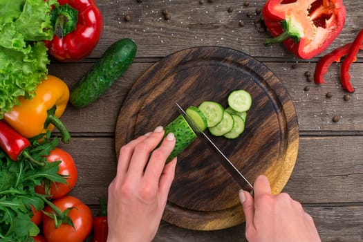 Female hands cutting cucumber at table, top view. On the table vegetables and a wooden board